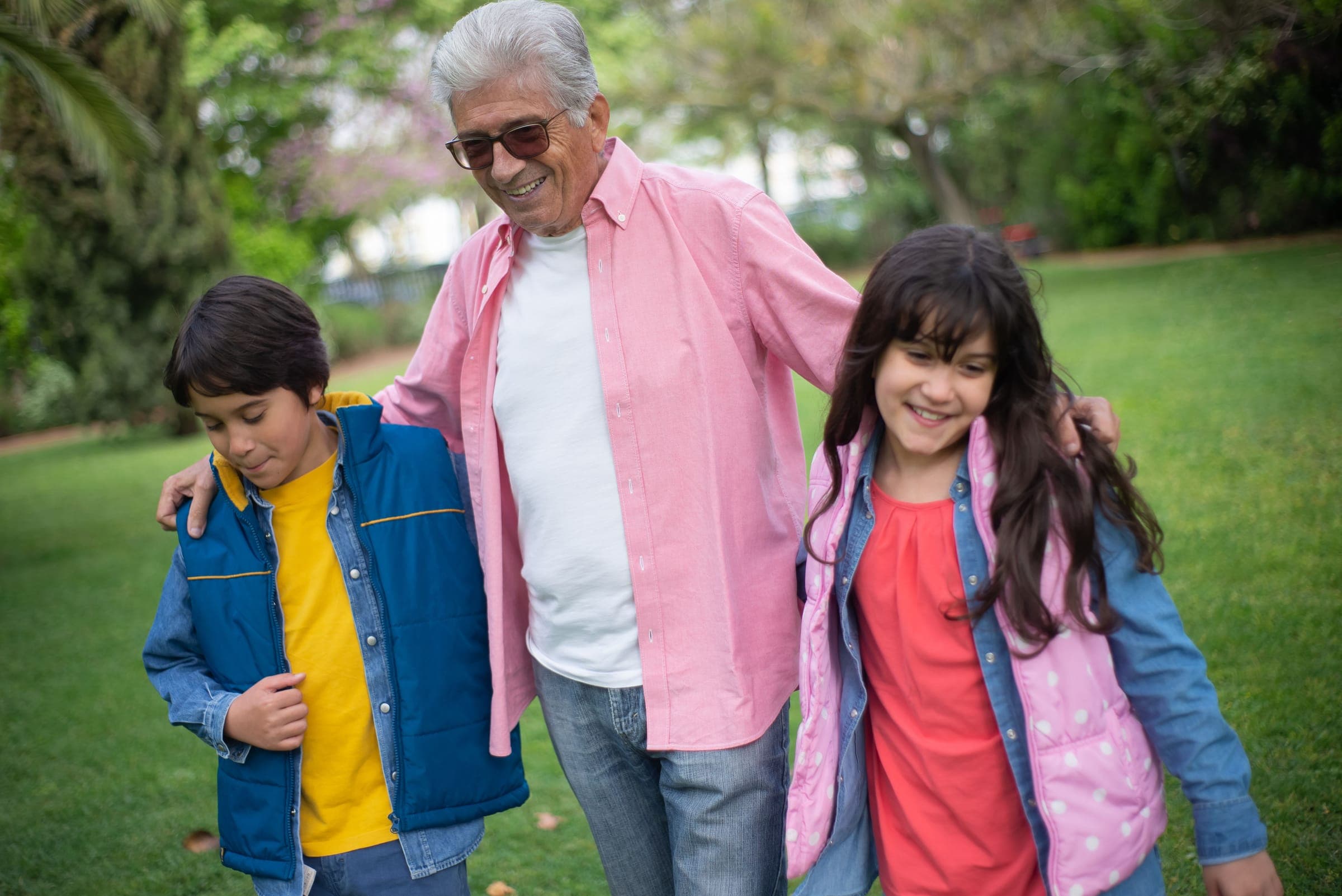 A smiling older man walks through a park with his arms around a boy and a girl