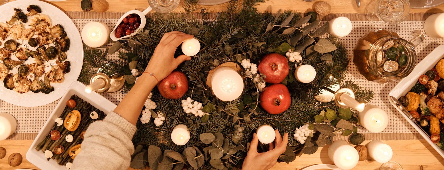 A holiday table seen from above, laid with candles, greenery and dishes of food, as a pair of hands sets the last candle in place