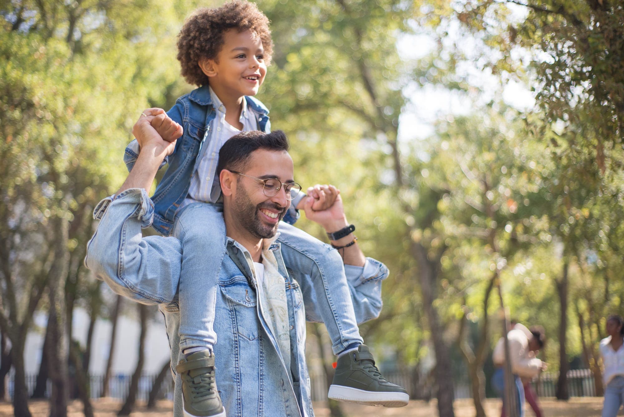 A laughing boy rides on a smiling man’s shoulders in a sunny park